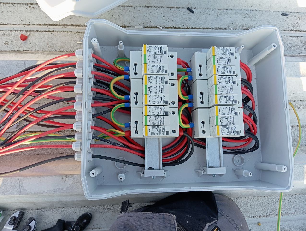 Otwarta skrzynka elektryczna z zabezpieczeniami przepięciowymi marki CITEL, liczne przewody w kolorach czarnym, czerwonym, zielono-żółtym, w tle fragment dachu i narzędzia.