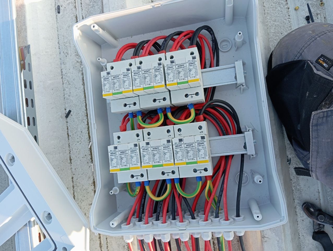 Otwarta skrzynka elektryczna z zainstalowanymi ogranicznikami przepięć CITEL, widoczne czerwone, czarne i żółto-zielone przewody, szara obudowa skrzynki i fragment dachu z elementami...