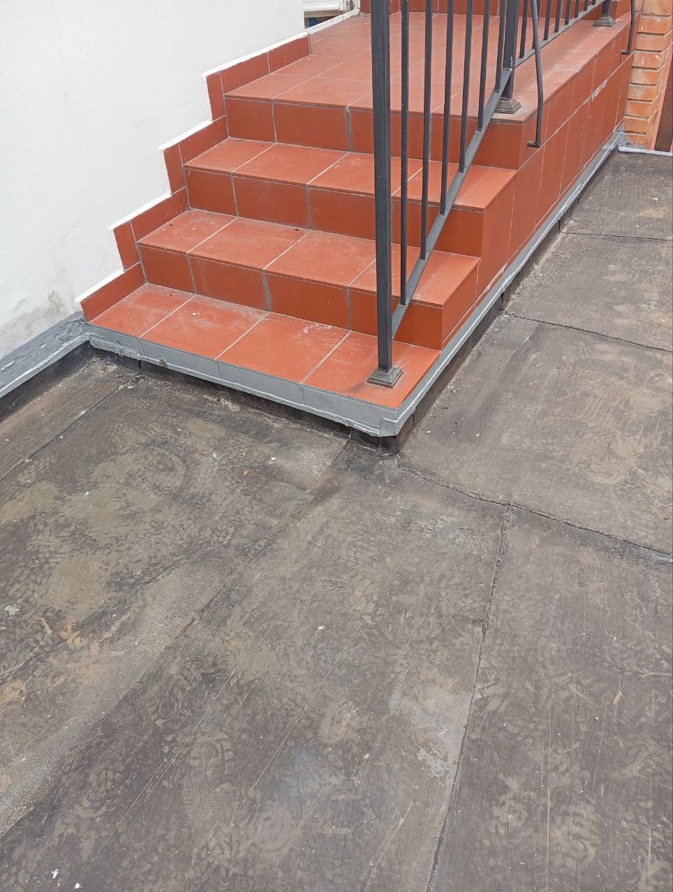 Wykończone schody zewnętrzne z terakoty w kolorze ceglastym, prowadzące na płaski dach pokryty bitumiczną papą, z widocznymi śladami obuwia.