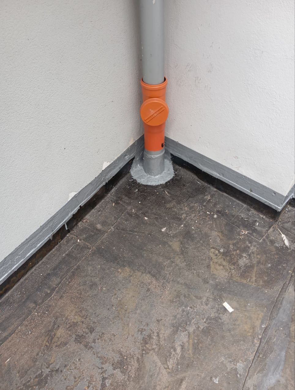 Szara rura kanalizacyjna z pomarańczowym zaworem, uszczelniona szarą masą przy podłodze w rogu pomieszczenia, widoczne ślady po malowaniu na ścianie i zabrudzona podłoga.