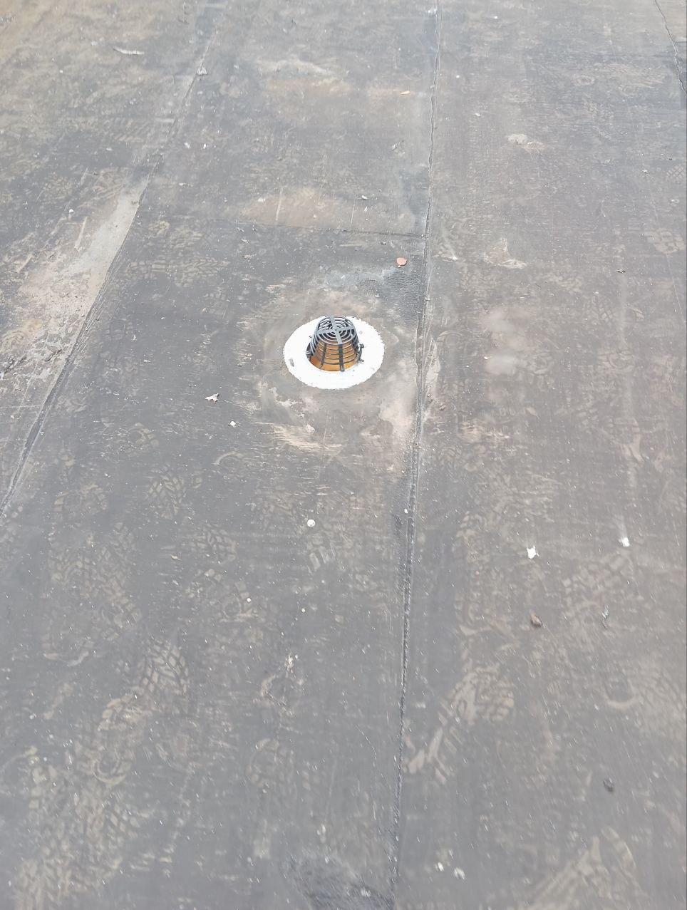 Widok z góry na płaski, bitumiczny dach z zamontowanym odpływem wody deszczowej zabezpieczonym kratką, pokryty śladami butów.