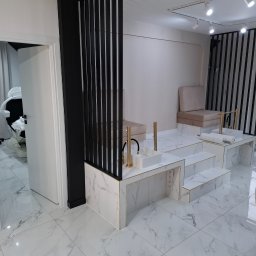 Firma Remontowo-Budowlana Władysław Szczechowicz - Nowoczesne wnętrze salonu kosmetycznego z podwyższonymi stanowiskami do manicure wykończonymi białym marmurem, złotymi detalami i czarnymi pionowymi listwami dekoracyjnymi, widok na otwarte drzwi...