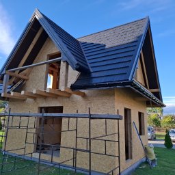 Firma Remontowo-Budowlana Władysław Szczechowicz - Dom w stanie surowym zamkniętym z czarną blachodachówką na dachu, widoczne rusztowania i pracownik na drabinie, w tle błękitne niebo.