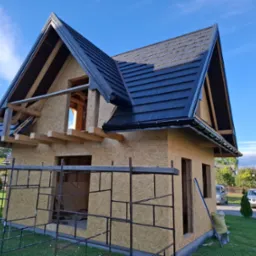 Dom w stanie surowym zamkniętym z czarną blachodachówką na dachu, widoczne rusztowania i pracownik na drabinie, w tle błękitne niebo.