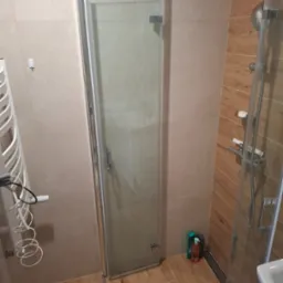 Zamknięta kabina prysznicowa ze szklanymi drzwiami w wykończonej łazience z beżowymi płytkami ściennymi i drewnopodobną podłogą, widoczny grzejnik drabinkowy i butelki kosmetyków w rogu.