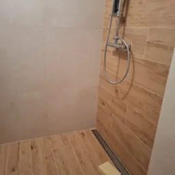 Nowoczesny prysznic walk-in z odpływem liniowym, wyłożony beżowymi płytkami imitującymi drewno na podłodze i ścianie, z chromowaną armaturą i beżową kostką mydła przy odpływie.