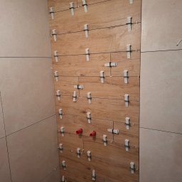 Firma Remontowo-Budowlana Władysław Szczechowicz - Ściana łazienki w trakcie układania płytek: połączenie płytek drewnopodobnych i beżowych, widoczne systemy poziomowania płytek, wystające rury instalacji wodnej.