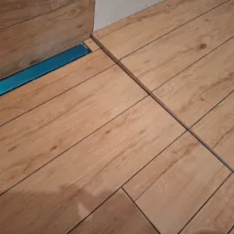 Ukośne ułożenie płytek podłogowych imitujących drewno, z widocznym liniowym odpływem prysznicowym z niebieskim elementem, podkreślającym nowoczesny charakter łazienki.