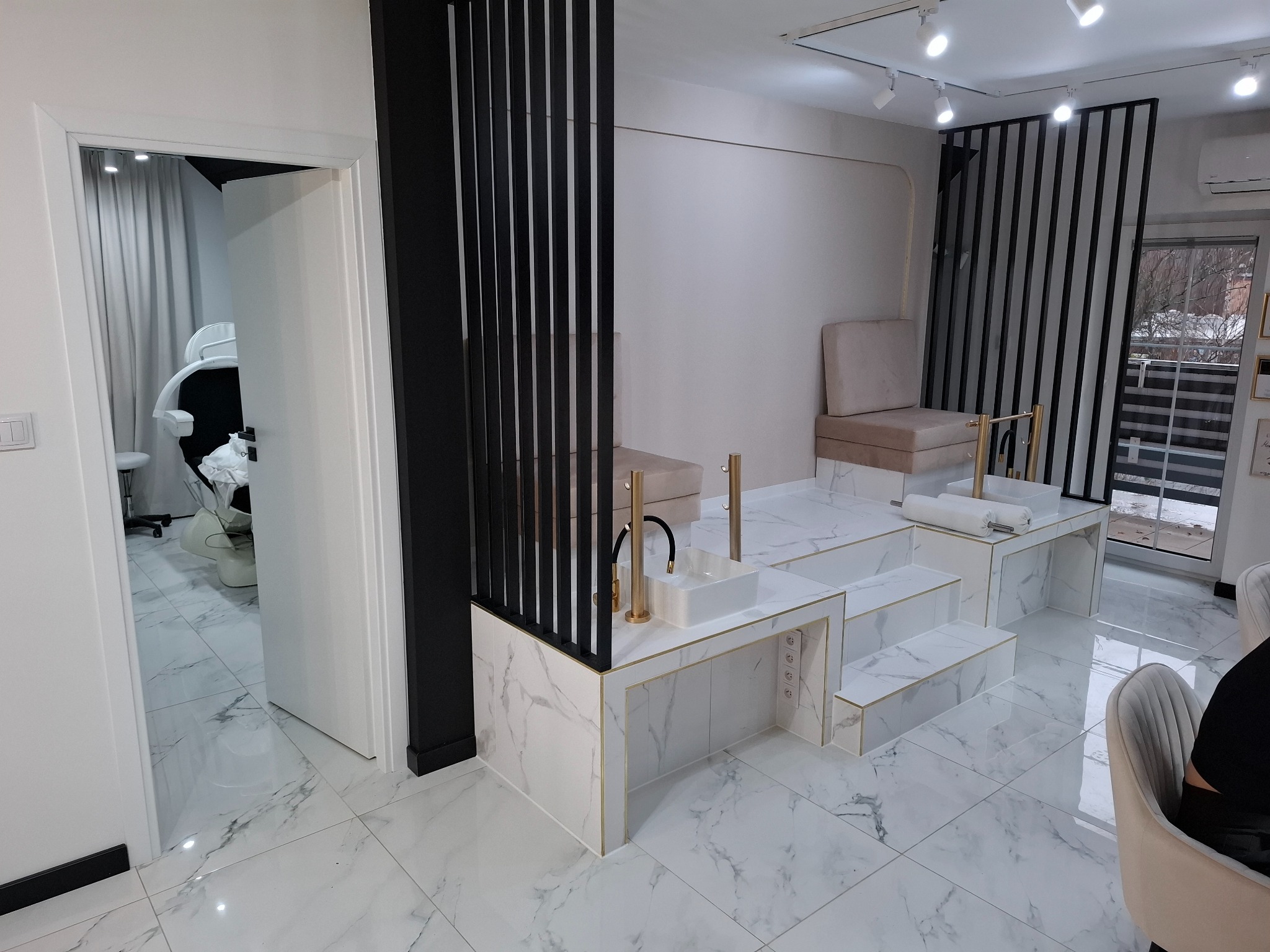 Nowoczesne wnętrze salonu kosmetycznego z podwyższonymi stanowiskami do manicure wykończonymi białym marmurem, złotymi detalami i czarnymi pionowymi listwami dekoracyjnymi, widok na otwarte drzwi...