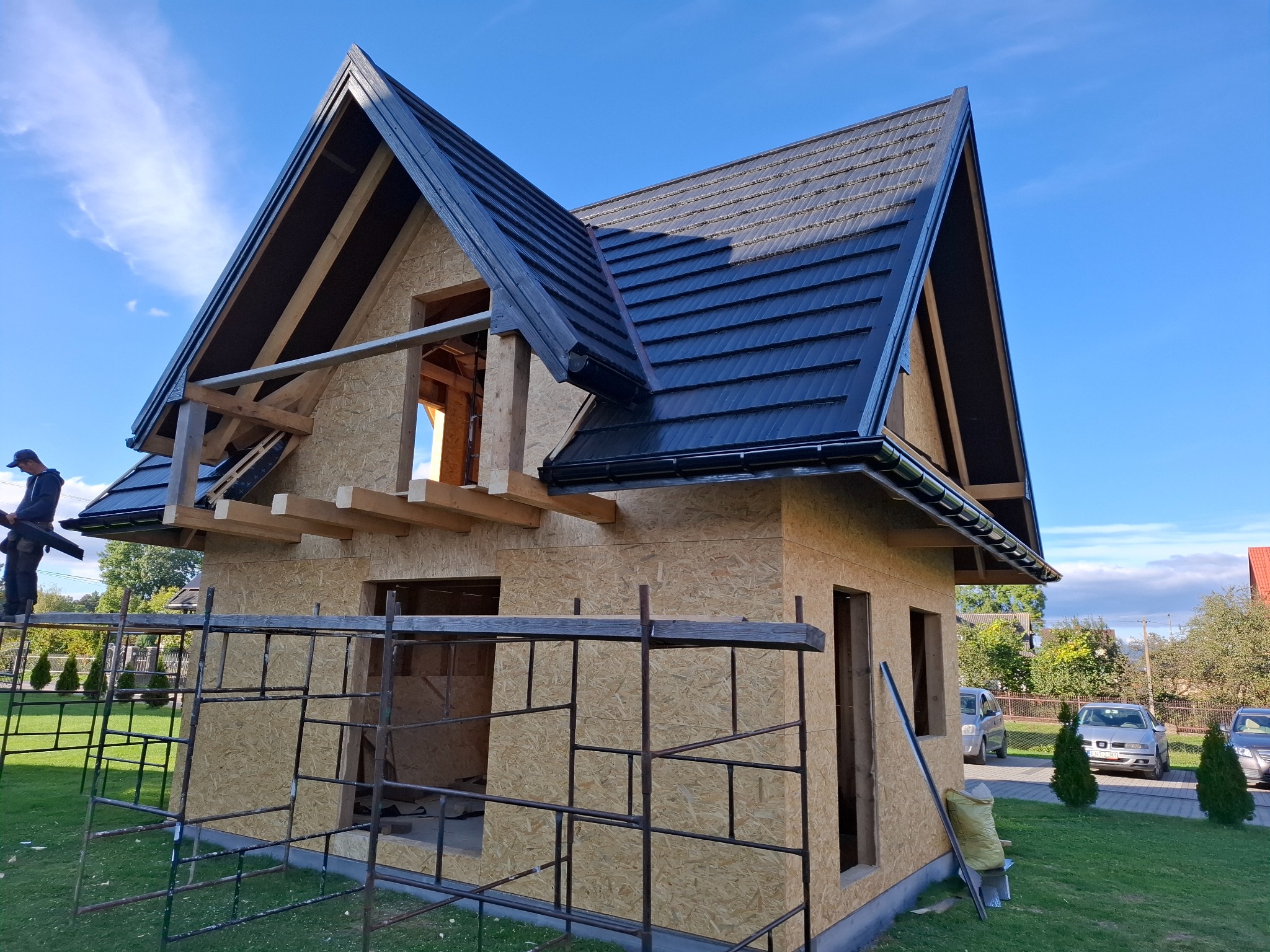 Dom w stanie surowym zamkniętym z czarną blachodachówką na dachu, widoczne rusztowania i pracownik na drabinie, w tle błękitne niebo.