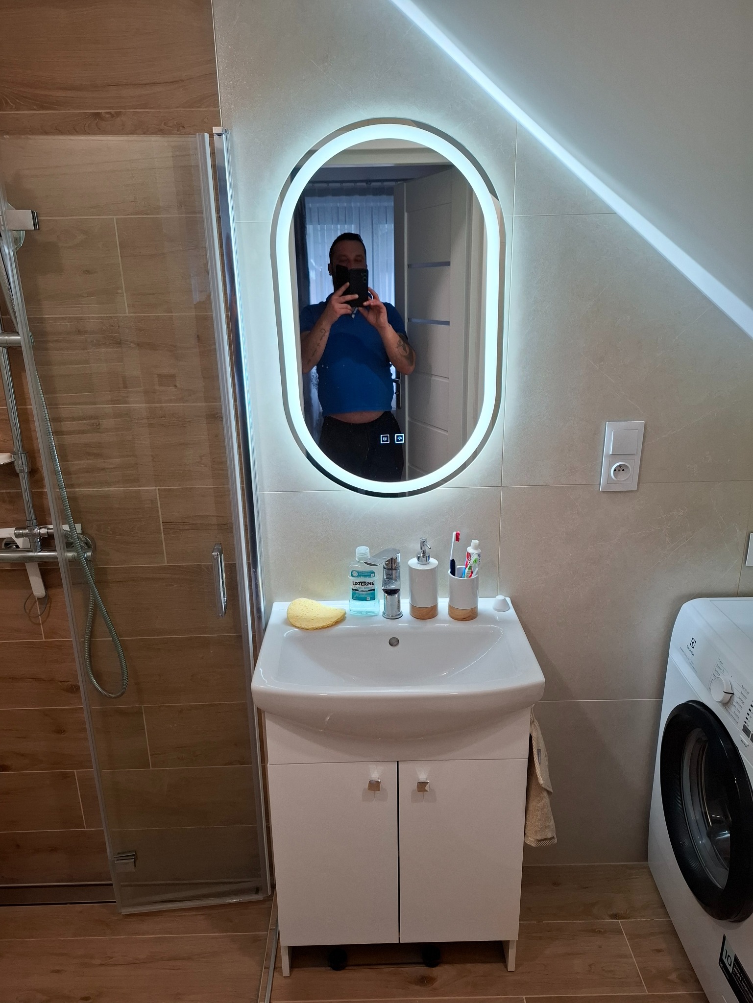 Nowoczesna łazienka z podświetlanym lustrem, umywalką z szafką i pralką, widoczny fragment kabiny prysznicowej z brązowymi płytkami imitującymi drewno oraz odbicie osoby robiącej zdjęcie w lustrze.