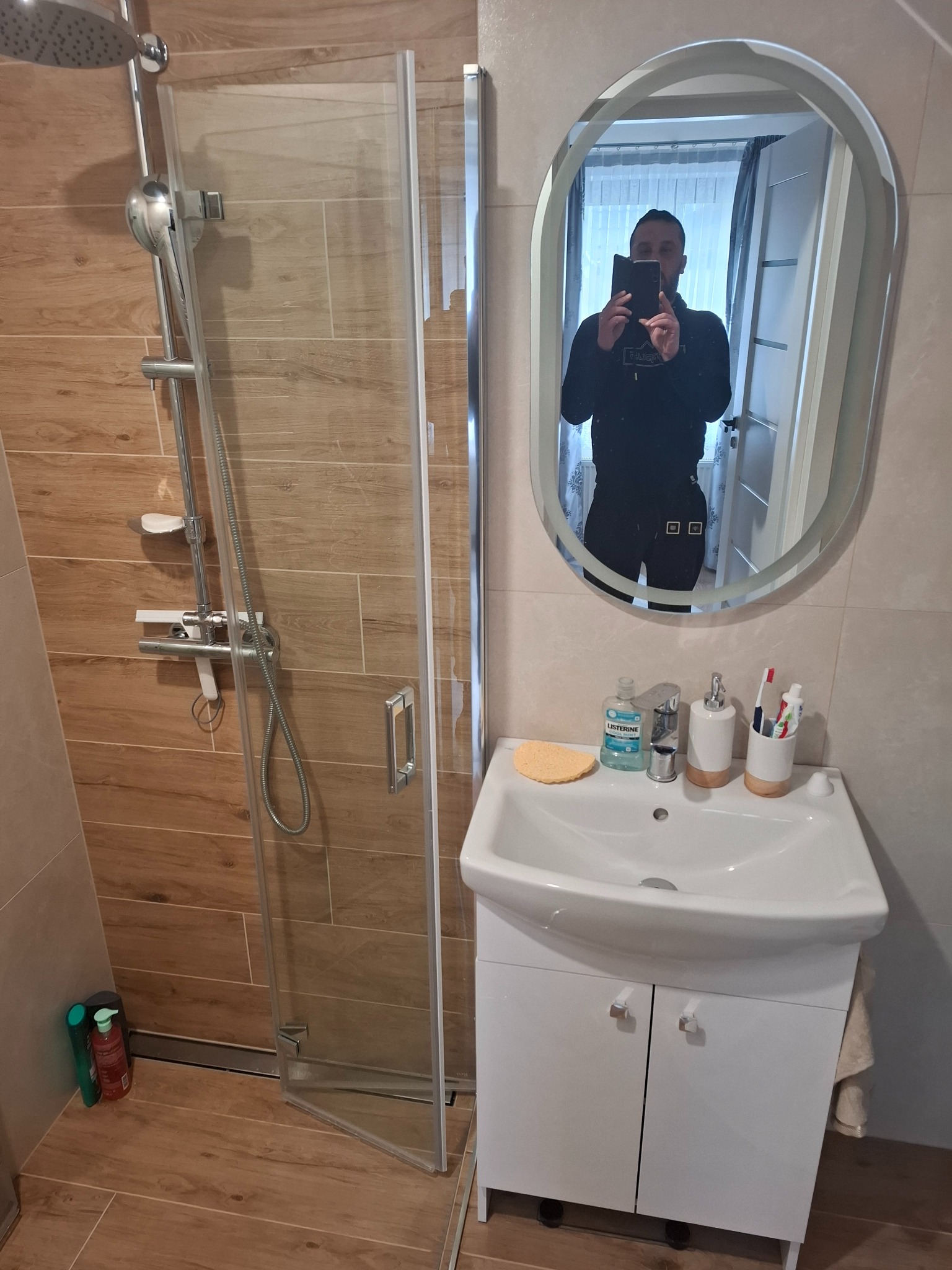 Nowoczesna łazienka z kabiną prysznicową typu walk-in, umywalką z szafką w kolorze białym oraz owalnym lustrem, w którym odbija się osoba robiąca zdjęcie.