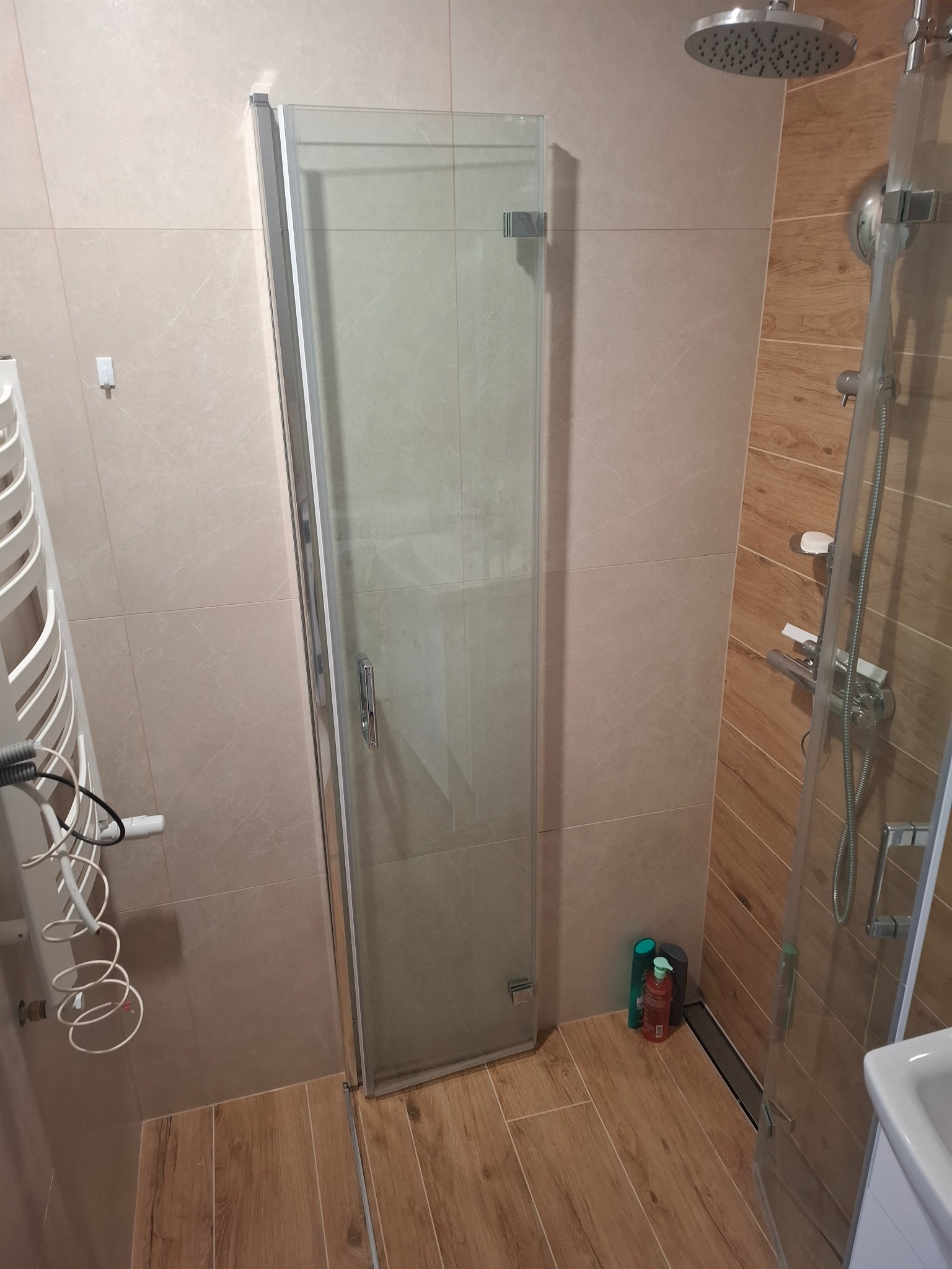 Zamknięta kabina prysznicowa ze szklanymi drzwiami w wykończonej łazience z beżowymi płytkami ściennymi i drewnopodobną podłogą, widoczny grzejnik drabinkowy i butelki kosmetyków w rogu.