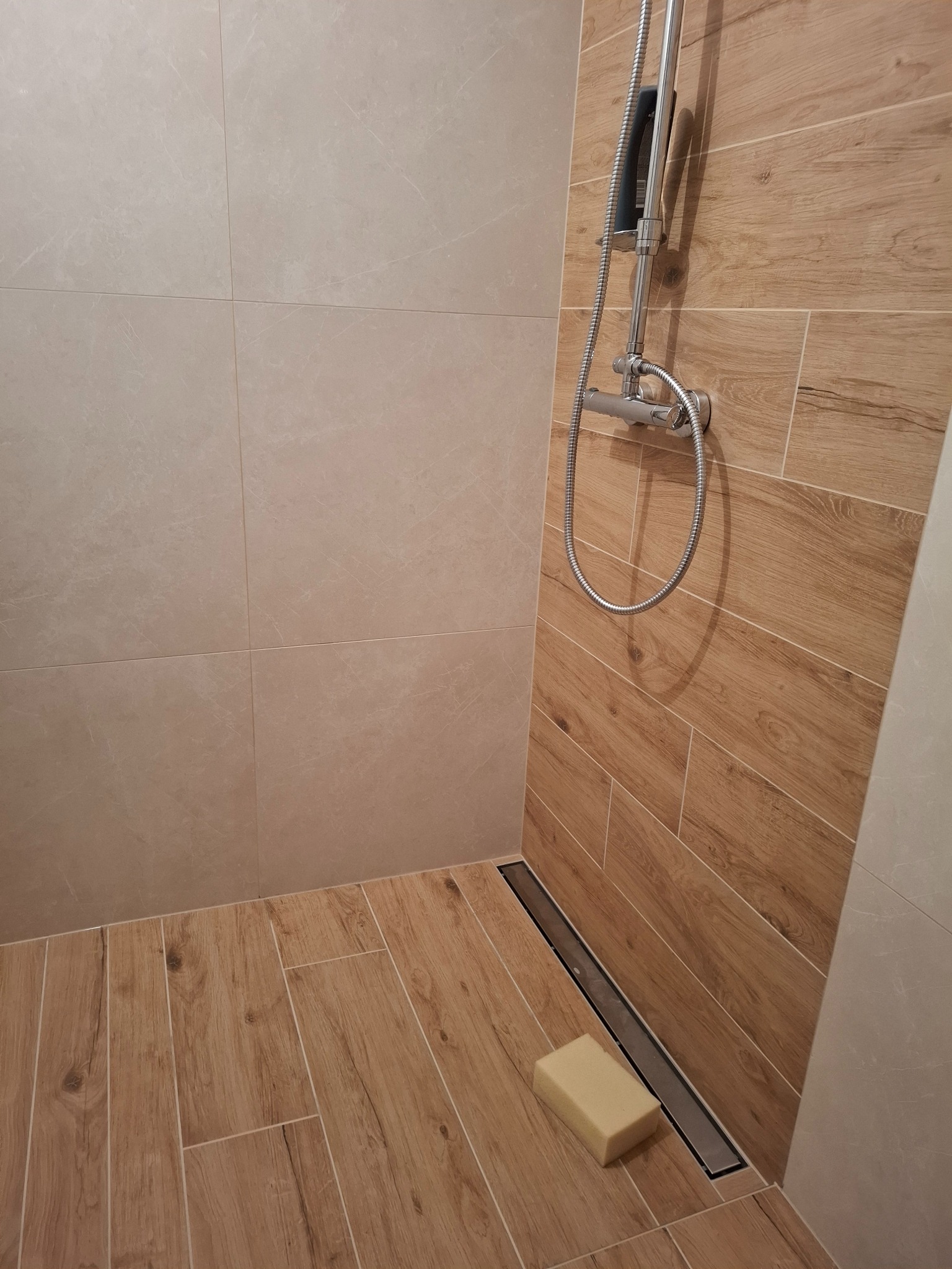 Nowoczesny prysznic walk-in z odpływem liniowym, wyłożony beżowymi płytkami imitującymi drewno na podłodze i ścianie, z chromowaną armaturą i beżową kostką mydła przy odpływie.