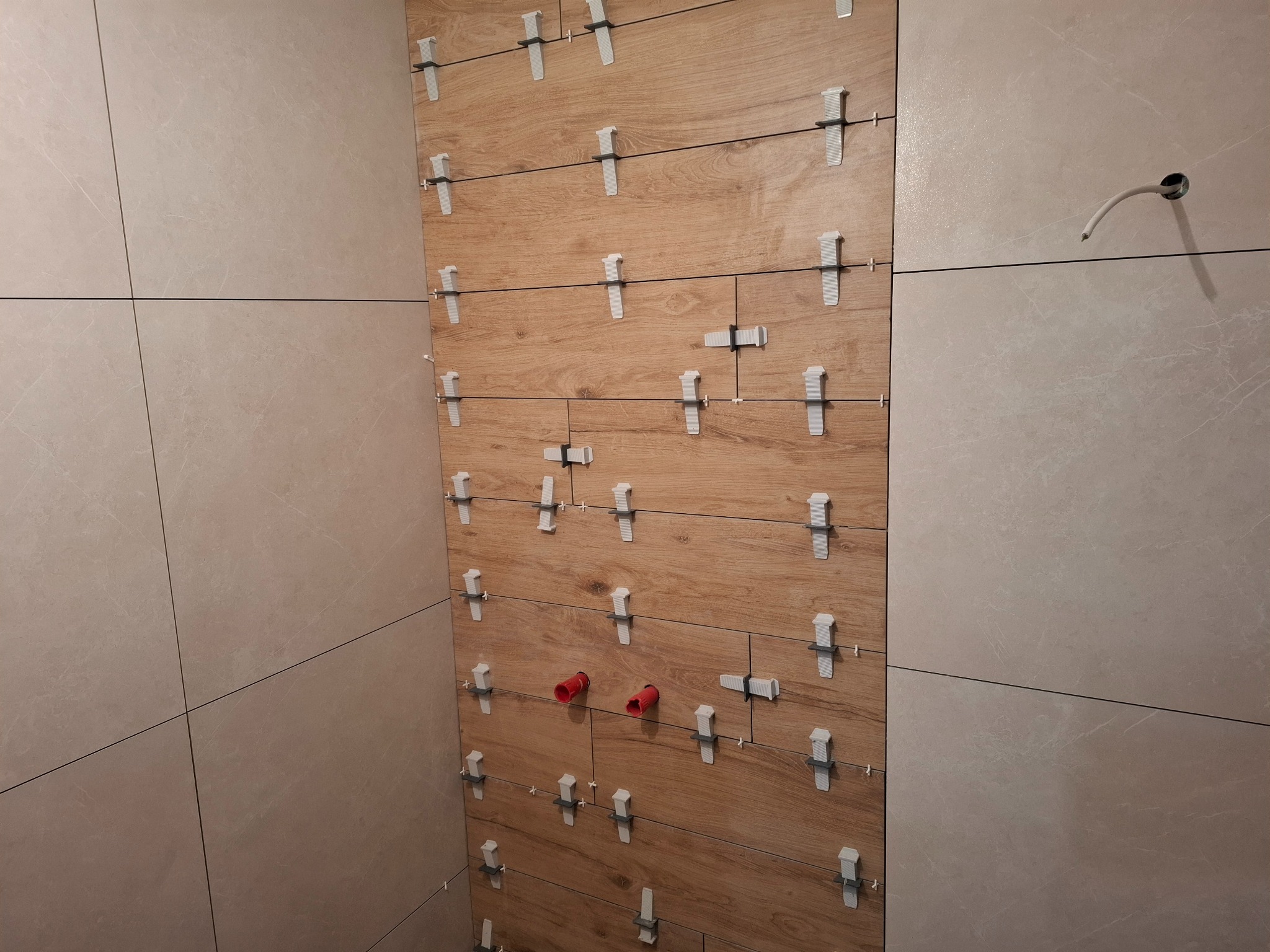 Ściana łazienki w trakcie układania płytek: połączenie płytek drewnopodobnych i beżowych, widoczne systemy poziomowania płytek, wystające rury instalacji wodnej.