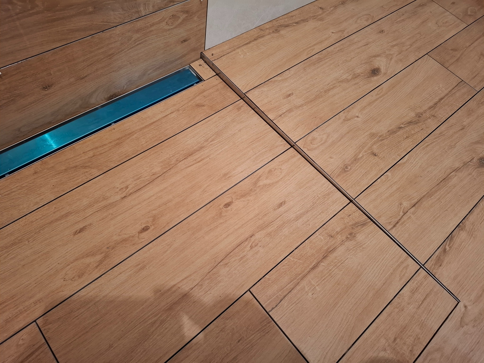 Ukośne ułożenie płytek podłogowych imitujących drewno, z widocznym liniowym odpływem prysznicowym z niebieskim elementem, podkreślającym nowoczesny charakter łazienki.