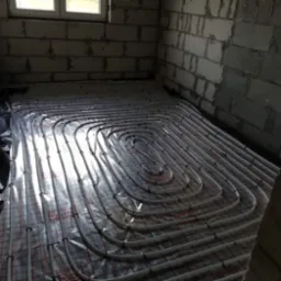 Instalacja ogrzewania podłogowego w surowym pomieszczeniu z bloczków, rury ułożone na folii z siatką, widoczne okno i fragment ściany z cegły.