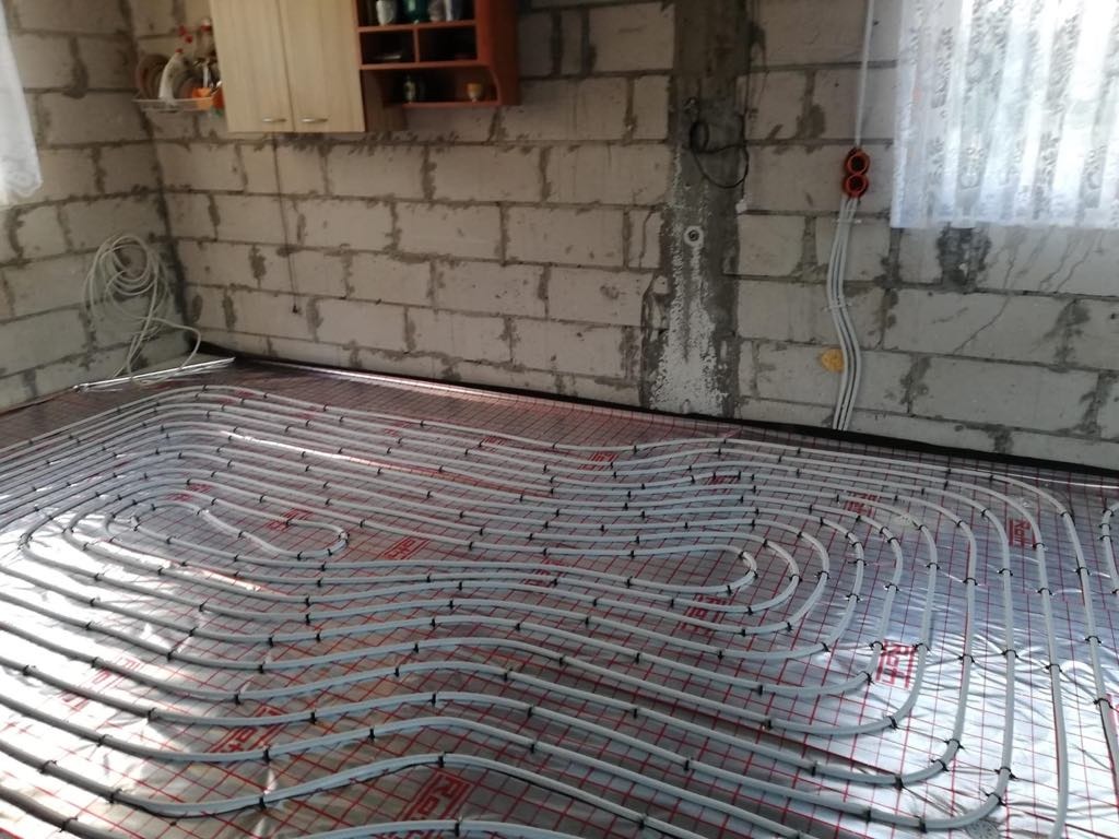 Instalacja ogrzewania podłogowego z rur ułożonych na folii izolacyjnej w surowym pomieszczeniu z betonowych bloczków i oknem z firanką.