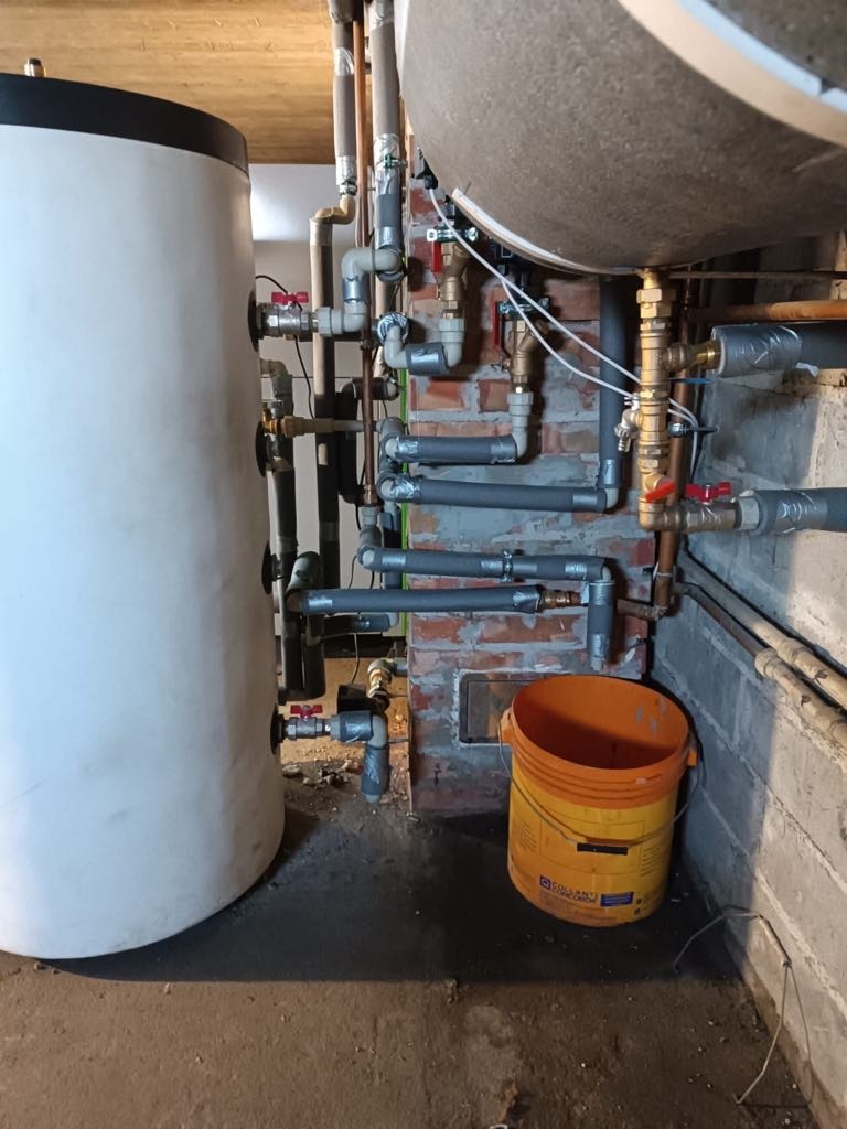 Złożona instalacja kotła na pellet z licznymi rurami miedzianymi i szarymi, zbiornik buforowy i zasobnik, widoczne elementy kotłowni w piwnicy.