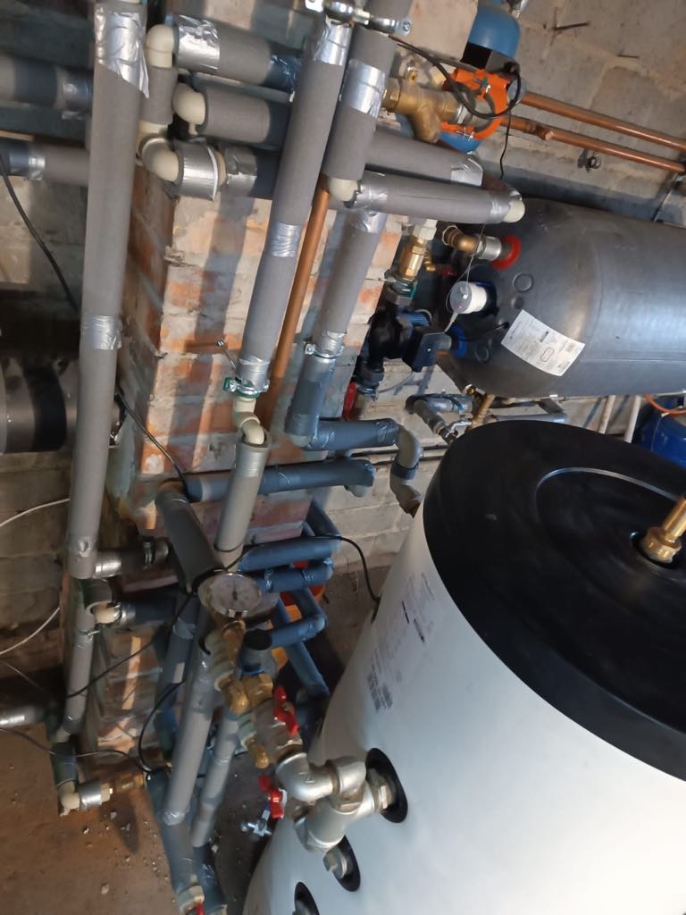 Złożona instalacja kotła na pellet z szarymi rurami izolowanymi taśmą, widoczny zbiornik i liczne zawory, całość na tle ceglanej ściany w kotłowni.