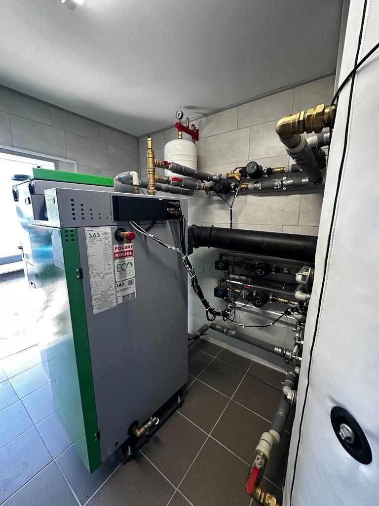 Szary kocioł SAS BIO EFFECT 12 KW z zielonym akcentem, zainstalowany w kotłowni z szarymi płytkami. Widoczne rury i elementy instalacji grzewczej.