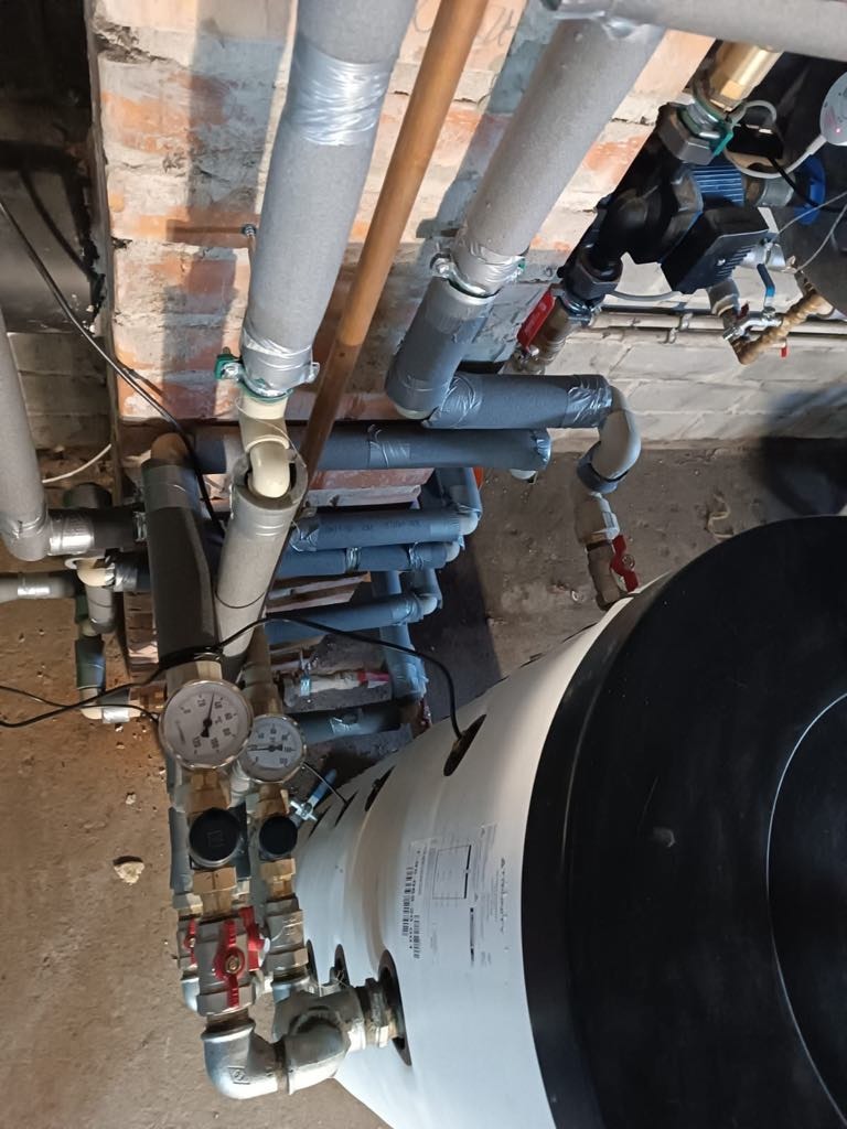 Instalacja kotła: biały zbiornik z manometrami, rury izolowane szarą pianką, zawory z czerwonymi pokrętłami. Widoczne połączenia hydrauliczne i ceglana ściana w tle.