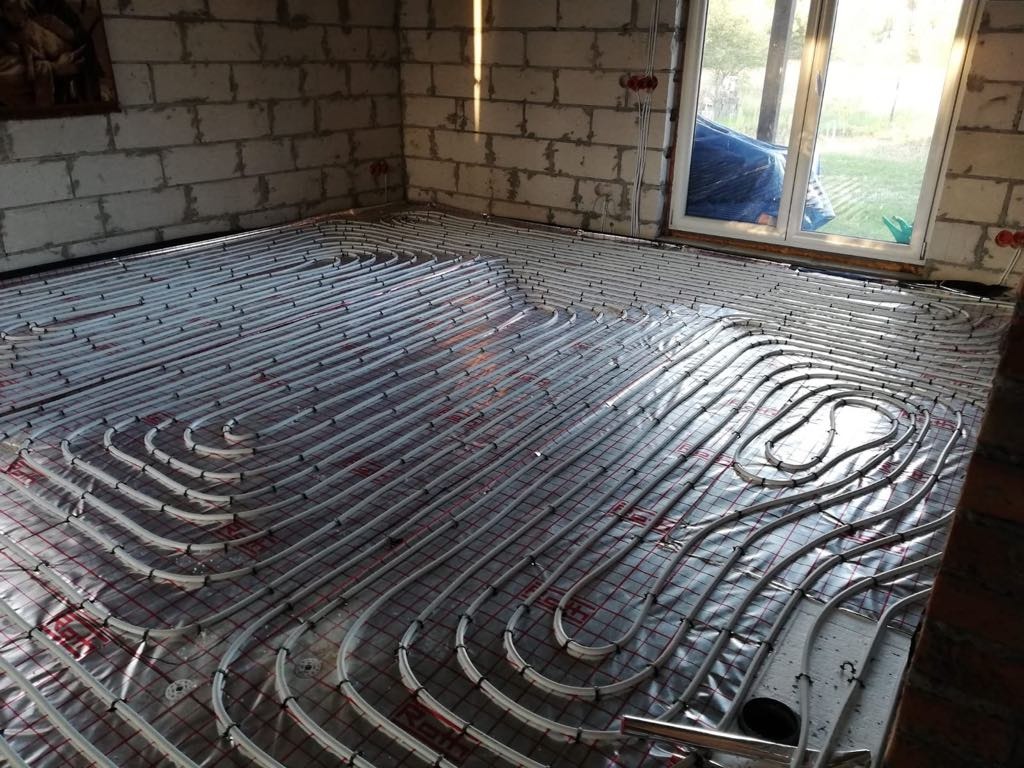 Białe rury ogrzewania podłogowego ułożone na folii w surowym wnętrzu. Widoczne okno i fragment ściany z bloczków. Instalacja przed zalaniem posadzką.