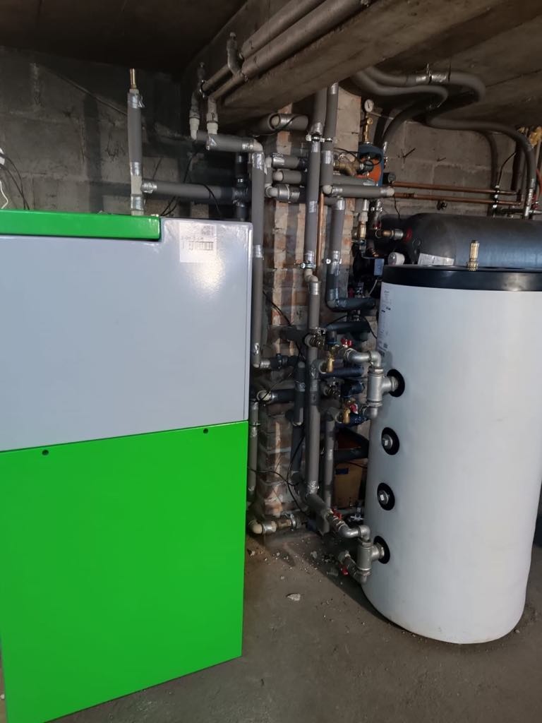 Zainstalowany kocioł na pellet SAS BIO EFFECT 14 kW w kotłowni. Widoczne rury instalacji CO i zasobnik ciepłej wody. Szare ściany, surowy betonowy strop.