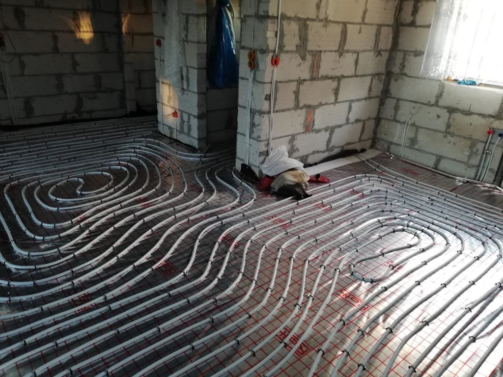Białe rury ogrzewania podłogowego ułożone na folii w surowym wnętrzu z szarych bloczków. Widoczne elementy instalacji i okno w tle.