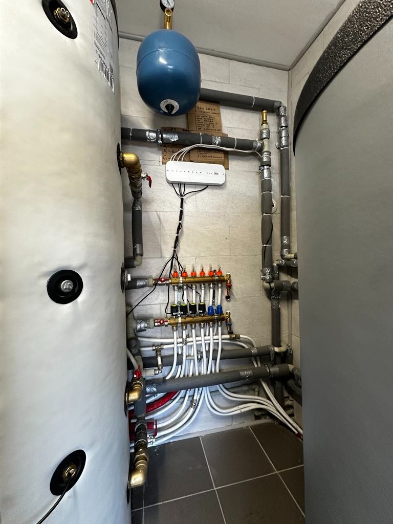 Kompletna instalacja hydrauliczna z zbiornikiem, rurami i rozdzielaczem, zamontowana w pomieszczeniu technicznym z szarą płytką podłogową i jasnymi ścianami.