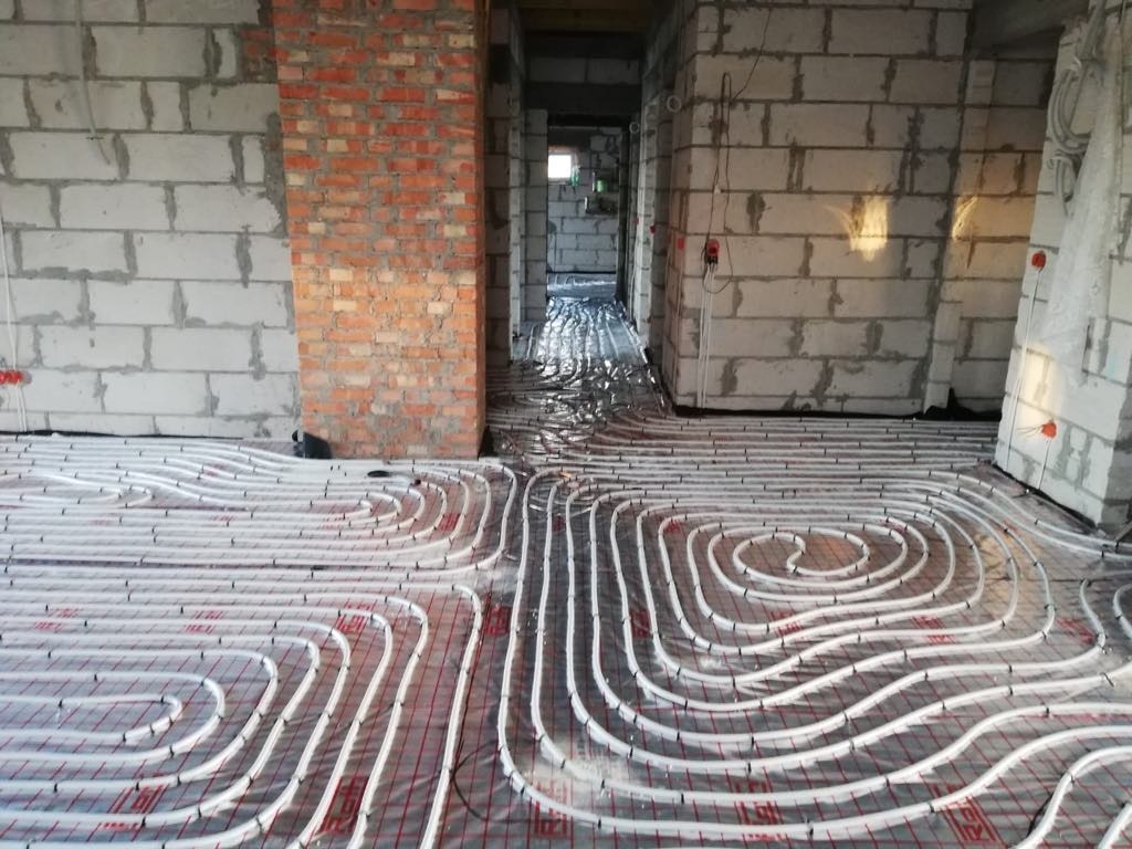 Białe rury ogrzewania podłogowego ułożone spiralnie na folii z czerwoną siatką, widoczne ściany z białego i czerwonej cegły w surowym stanie.