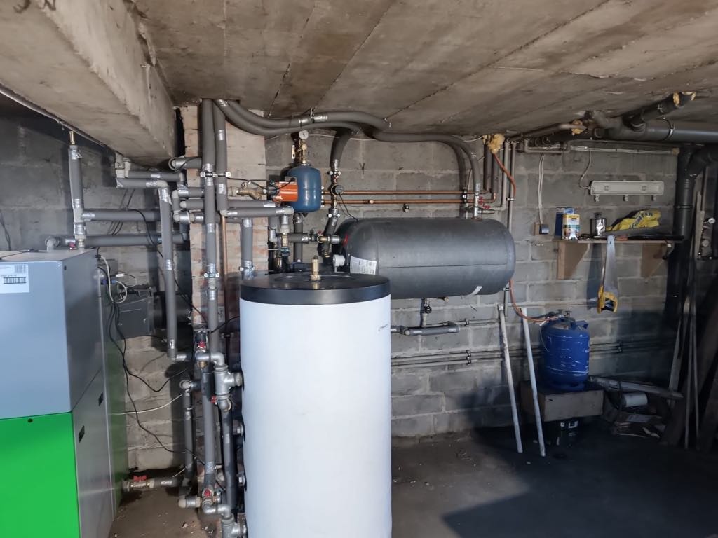 Kocioł na pellet SAS BIO EFFECT 14 kW: instalacja w kotłowni z szarą ścianą z cegły, widoczne rury, zbiornik buforowy i elementy instalacji grzewczej.