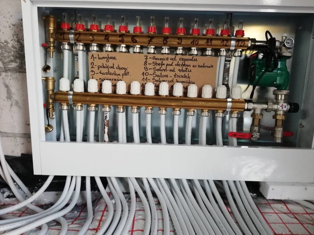 Rozdzielacz ogrzewania podłogowego w białej szafce z opisem stref na kartce. Widoczne rury i zawory. Instalacja grzewcza z pompą obiegową.