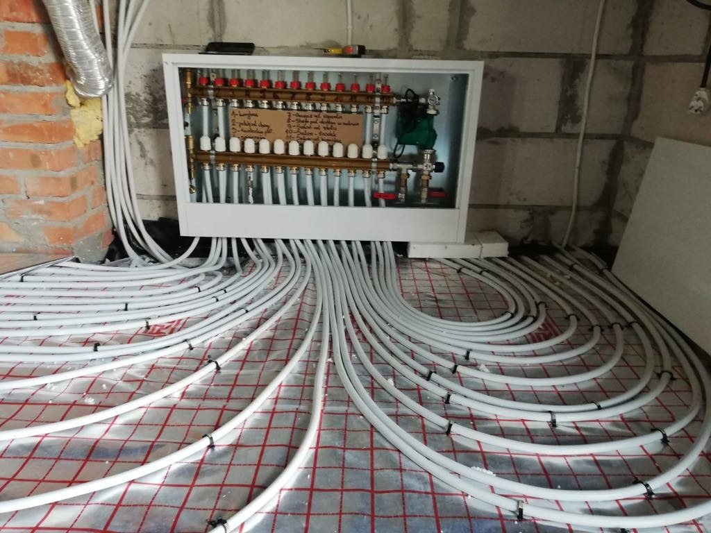 Rozdzielacz ogrzewania podłogowego z białymi rurami ułożonymi na folii z czerwoną kratką, widoczne przyłącza i ściany w surowym stanie.