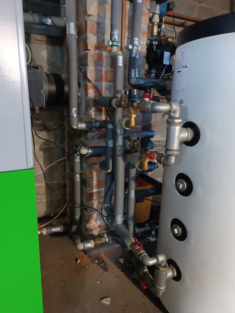 Instalacja kotła pellet: rury z izolacją, zawory, pompa obiegowa, zbiornik buforowy. Widoczny fragment kotła z zielonym panelem bocznym, ściana z cegły w tle.