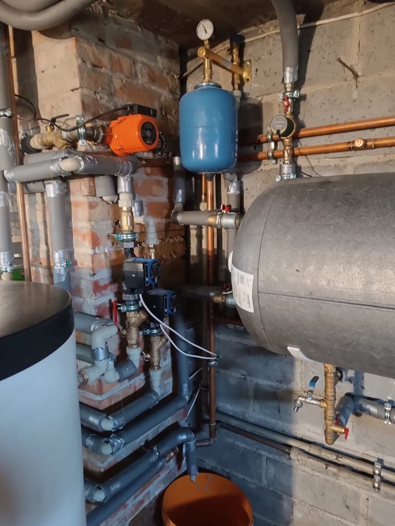 Złożona instalacja kotła: rury miedziane i plastikowe, zbiornik szary i niebieski, pompy obiegowe (pomarańczowa i czarna), manometr, zawory, ceglany fragment ściany.