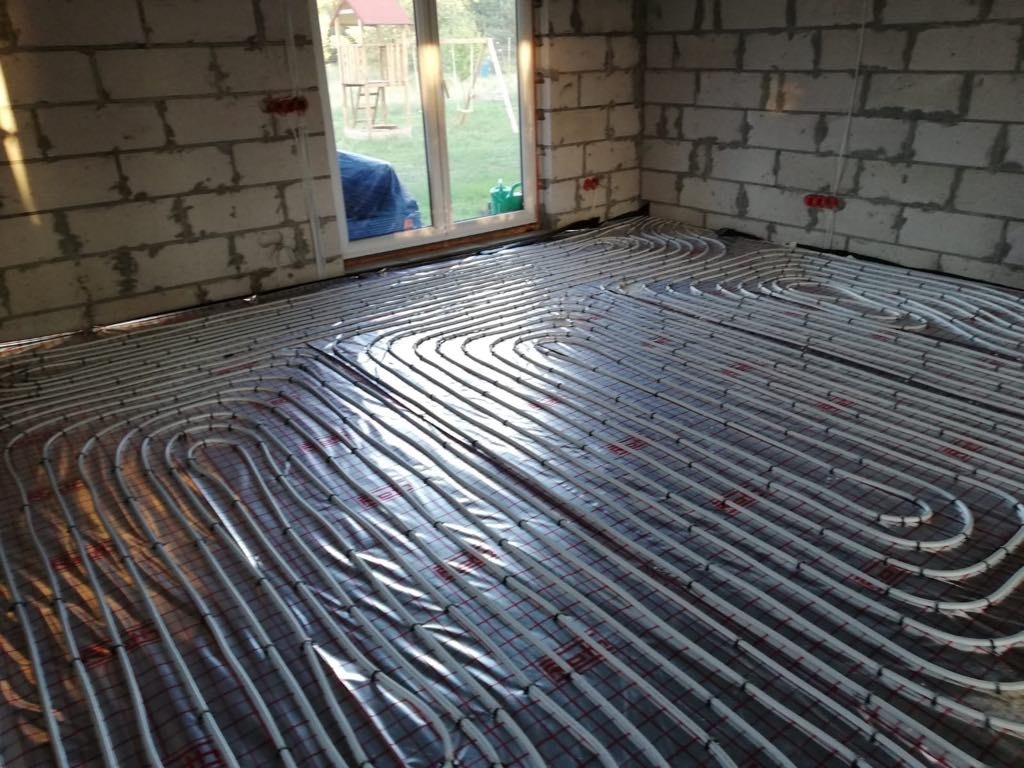 Białe rury ogrzewania podłogowego ułożone na folii izolacyjnej w surowym wnętrzu z oknem na zewnątrz. Widoczne ściany z bloczków betonowych.