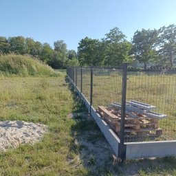 Tomasz Malarski - Montaż ogrodzenia panelowego na betonowej podmurówce. Widoczne elementy ogrodzenia na palecie. Trawa i drzewa w tle. Słoneczny dzień.