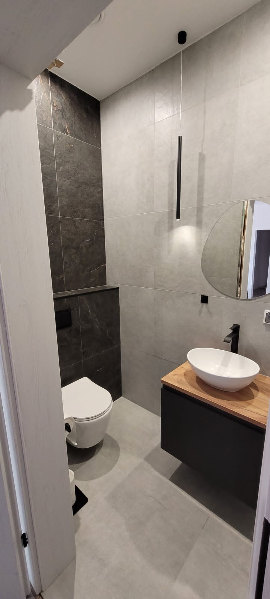 Nowoczesna łazienka z szarymi i czarnymi płytkami, umywalką na drewnianym blacie i wiszącą toaletą. Minimalistyczna lampa wisząca. Widok na fragment otwartych drzwi.