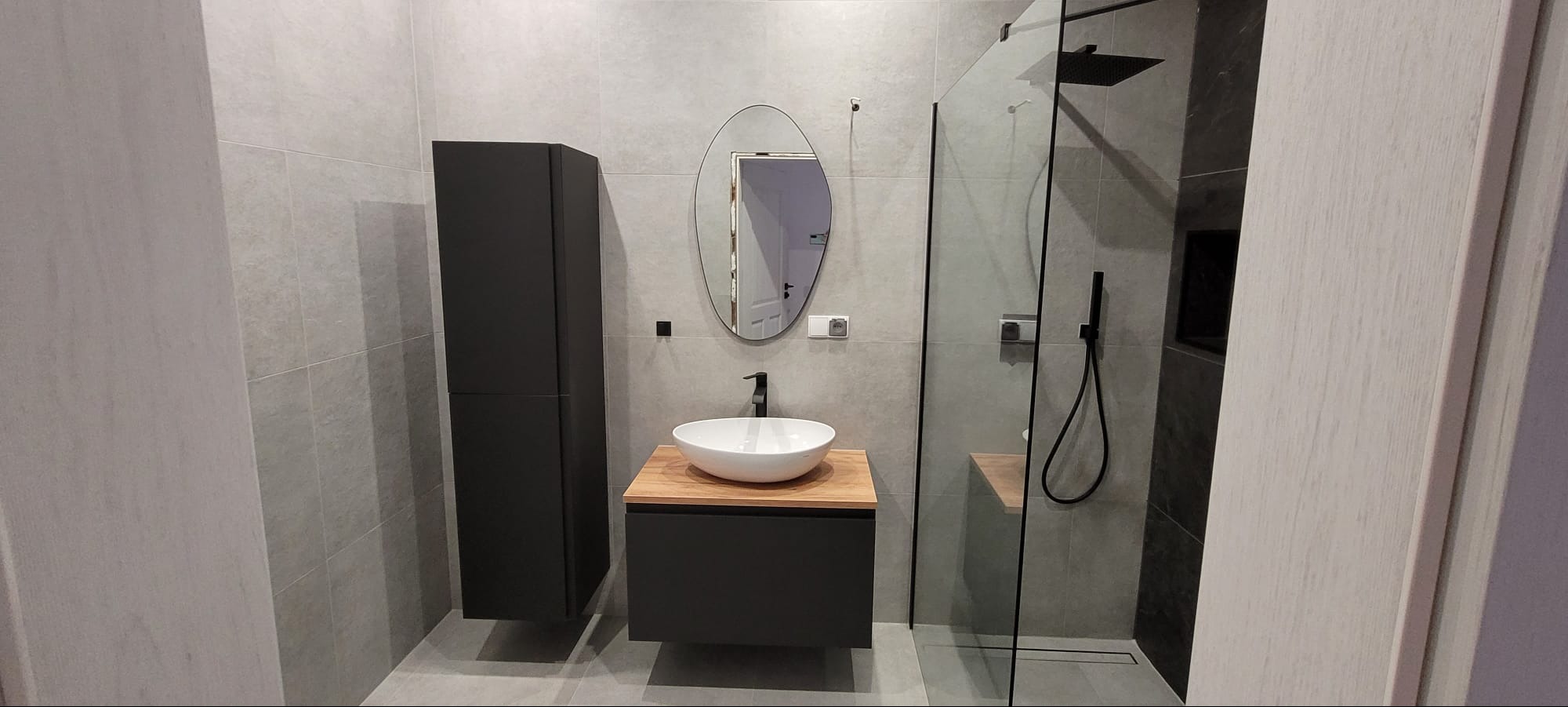 Nowoczesna łazienka z szarymi płytkami, czarnymi szafkami i drewnianym blatem pod umywalką. Kabina prysznicowa ze szklaną ścianką i czarną armaturą.