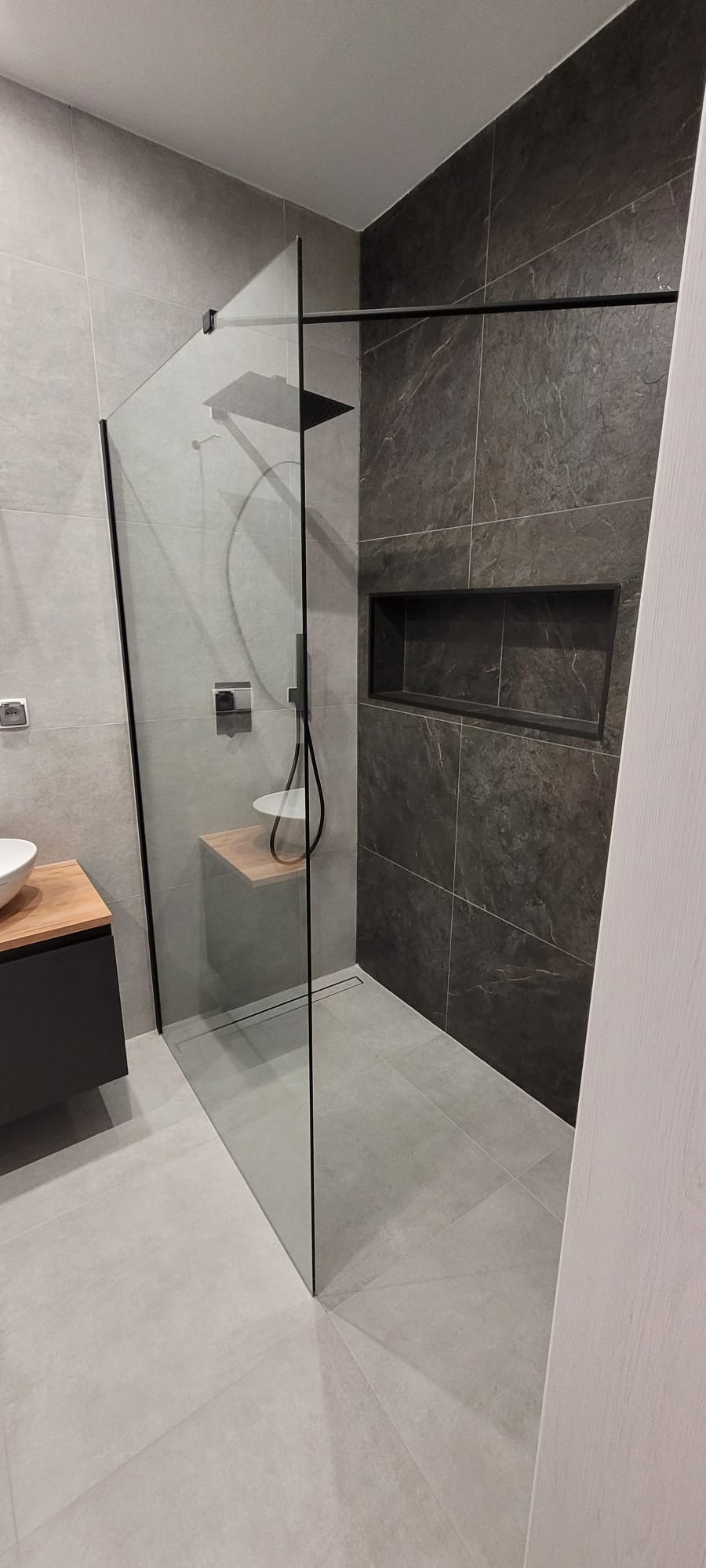 Nowoczesna kabina prysznicowa typu walk-in z czarnymi detalami i półką z drewna. Ciemne płytki na ścianie z wnęką dodają elegancji wnętrzu.