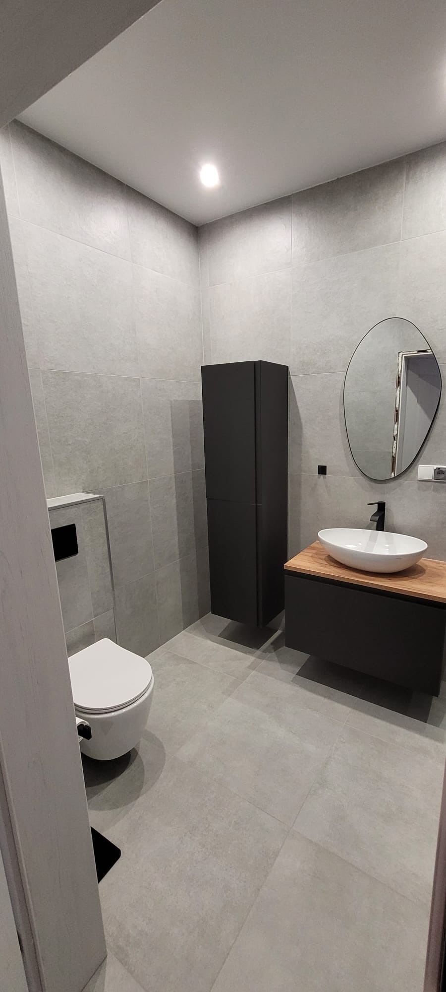 Nowoczesna, szara łazienka po remoncie z owalnym lustrem, wiszącą szafką i umywalką na drewnianym blacie, z widoczną toaletą.
