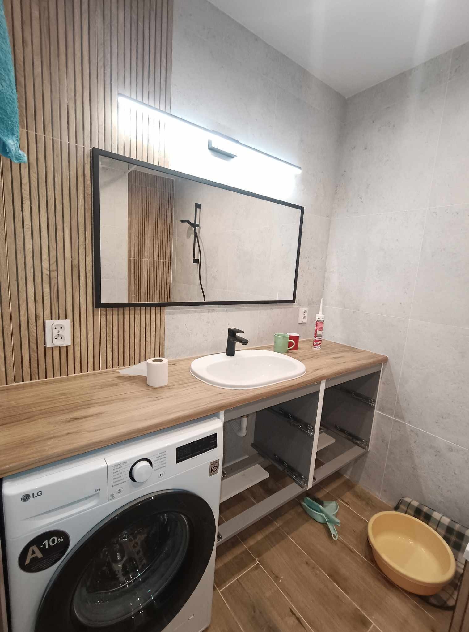 Nowoczesna łazienka z drewnianymi panelami, umywalką nablatową, lustrem w czarnej ramie i pralką LG. Widoczne płytki w odcieniach szarości i drewniana podłoga.
