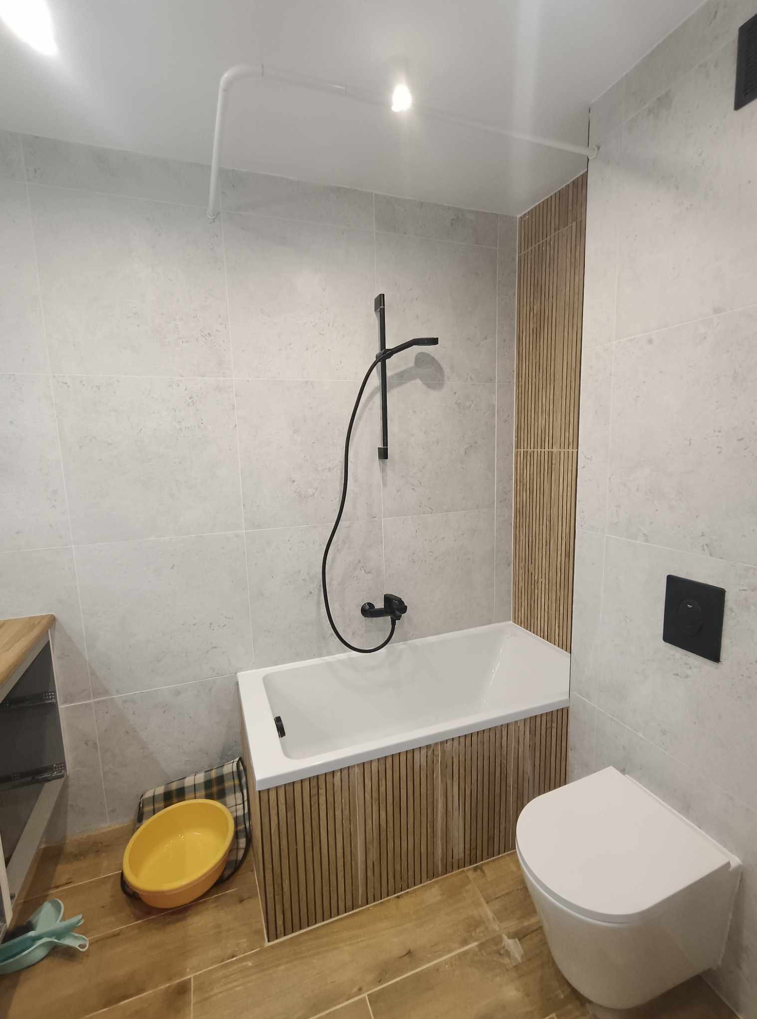 Biała wanna z czarną armaturą i obudową z drewnianych listewek w wykończonej łazience. Na podłodze płytki imitujące drewno i żółta miska. Szare płytki na ścianach.