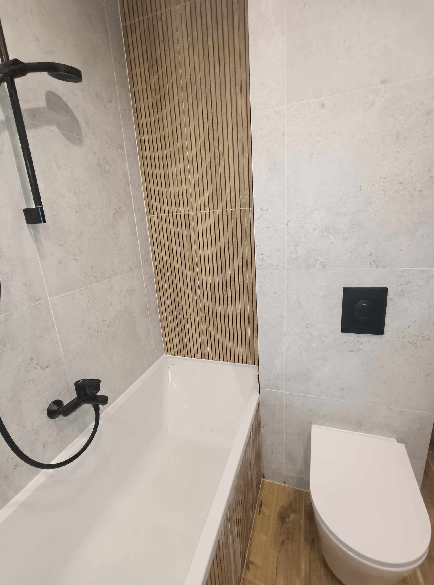 Nowoczesna łazienka po remoncie: biała wanna z czarną armaturą, toaleta w zabudowie, ściany w jasnych płytkach i dekoracyjne panele drewniane.