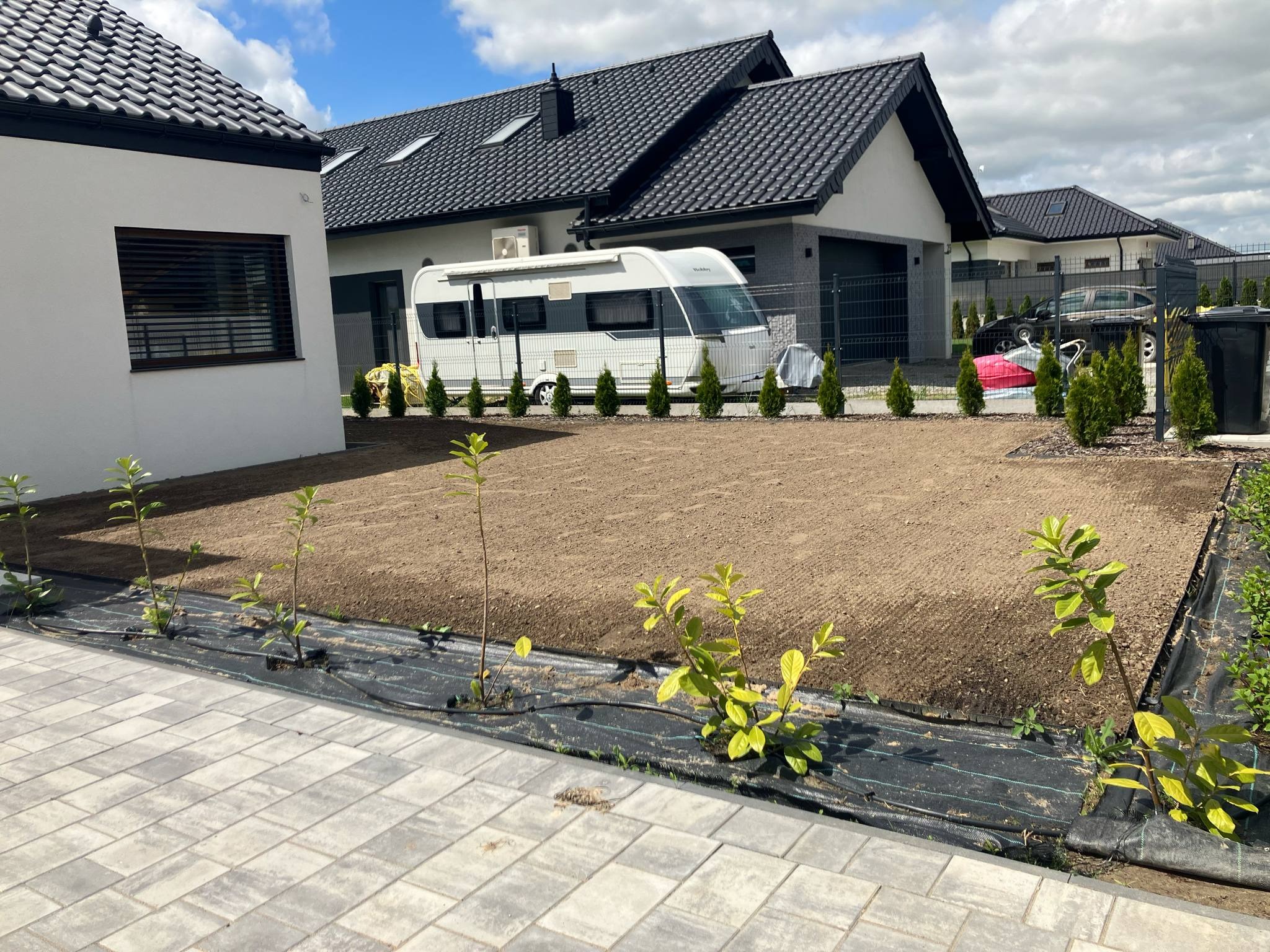 Przygotowany teren pod trawnik z rolki, widoczne młode drzewka wzdłuż obrzeża, domy w tle i przyczepa kempingowa. Grunt wyrównany, gotowy do położenia trawy.