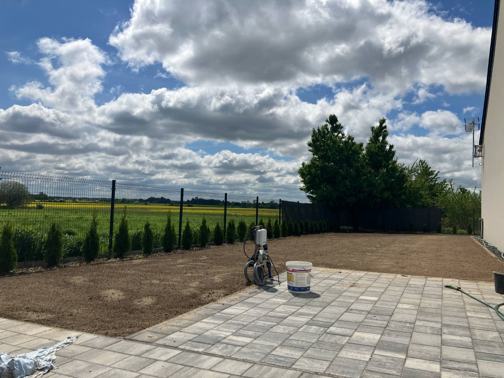 Przygotowany teren pod trawnik z widocznym systemem nawadniania, obrzeżami z tuj oraz ogrodzeniem panelowym. Ułożona kostka brukowa i malownicze chmury na niebie.