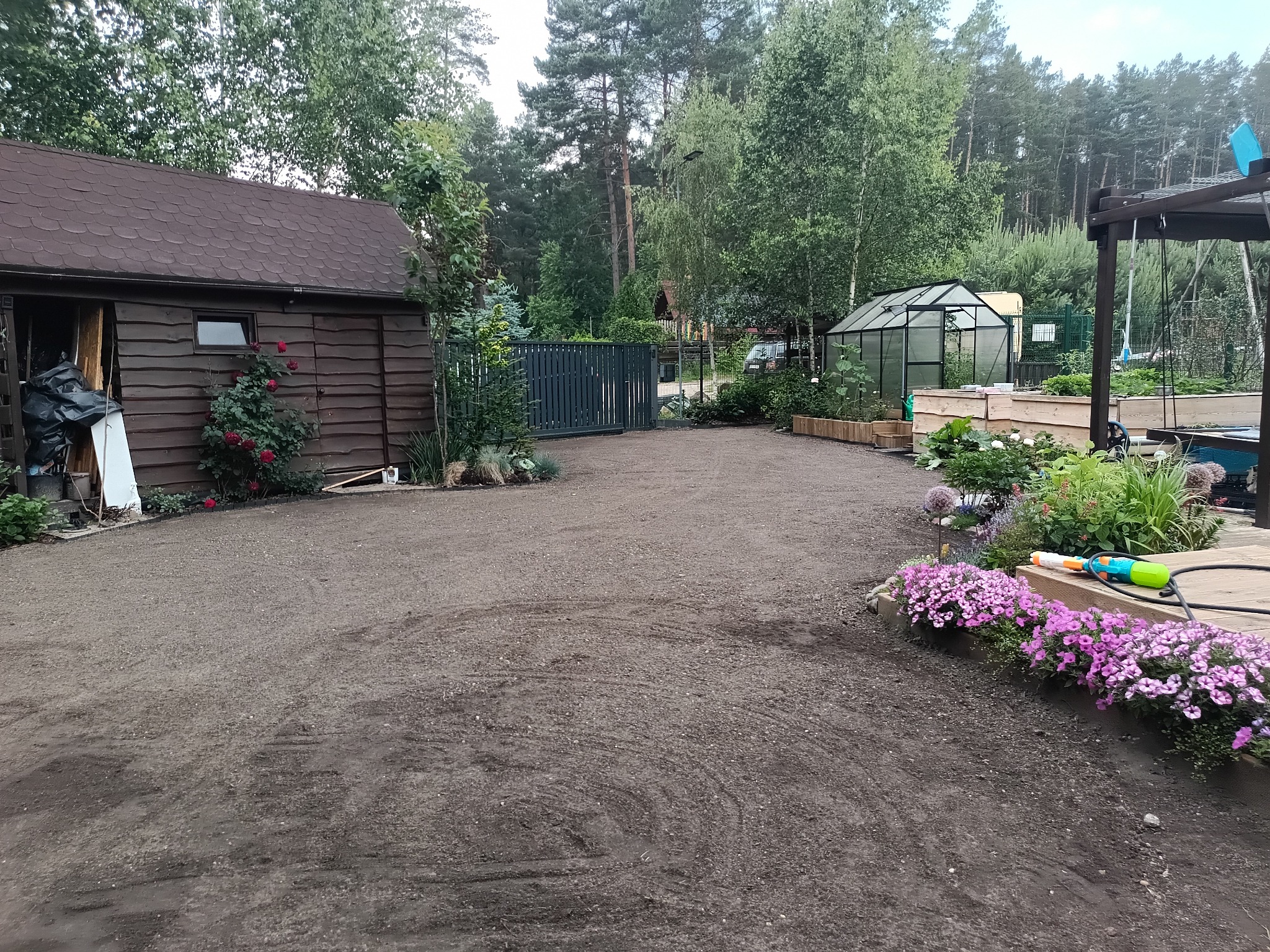 Przygotowany teren pod trawnik z widocznym domkiem narzędziowym, rabatami kwiatowymi i szklarnią w tle. Widoczna struktura ziemi po niwelacji.
