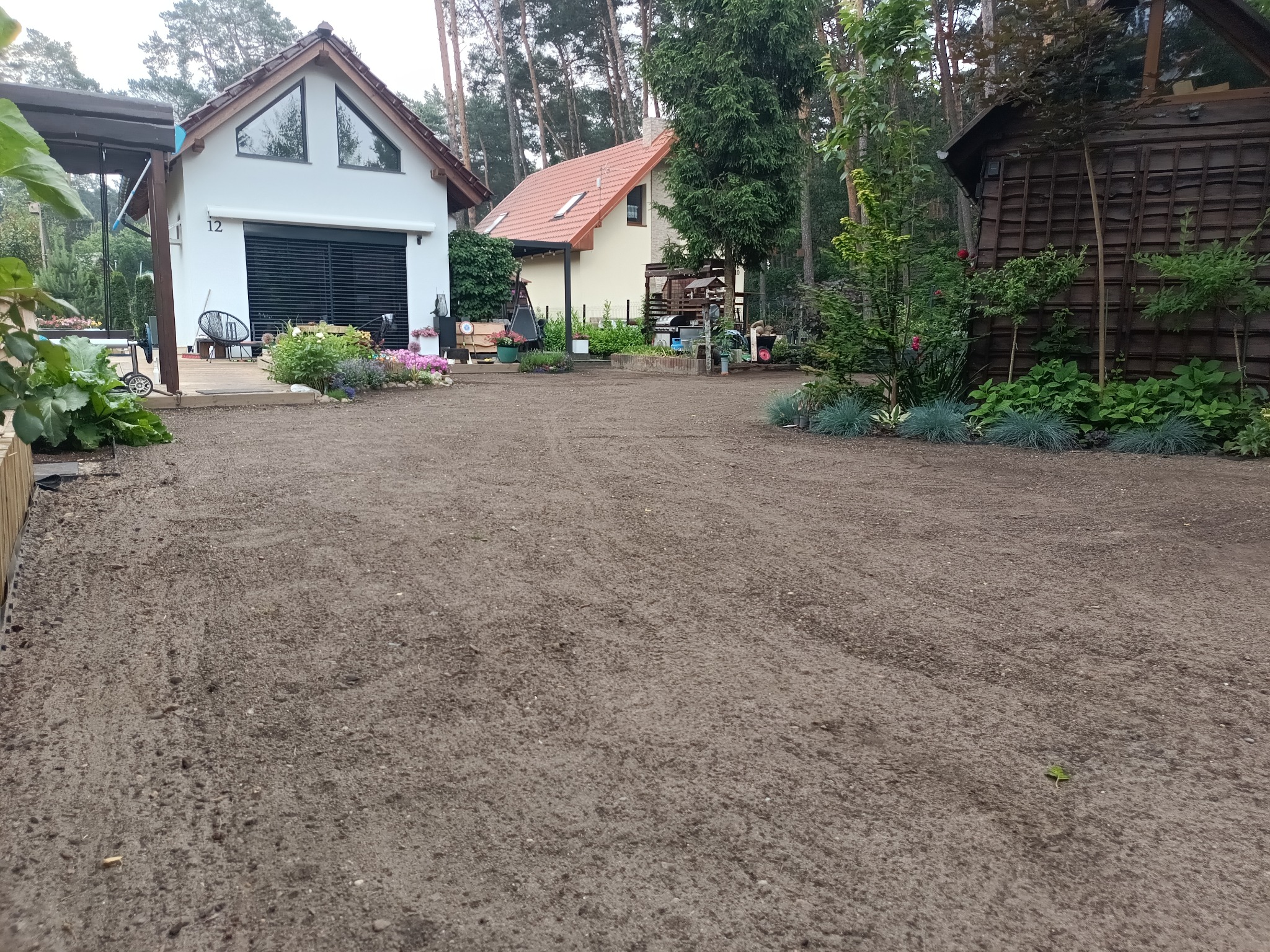 Przygotowany teren pod trawnik z rolki, widoczne budynki w tle, drzewa i rośliny. Teren wyrównany, gotowy do ułożenia trawy.