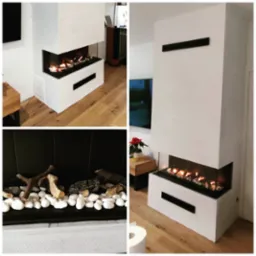 Nowoczesny, narożny wkład z płomieniami, obudowany białą konstrukcją z czarnymi akcentami, wykończony białymi kamieniami i drewnem.